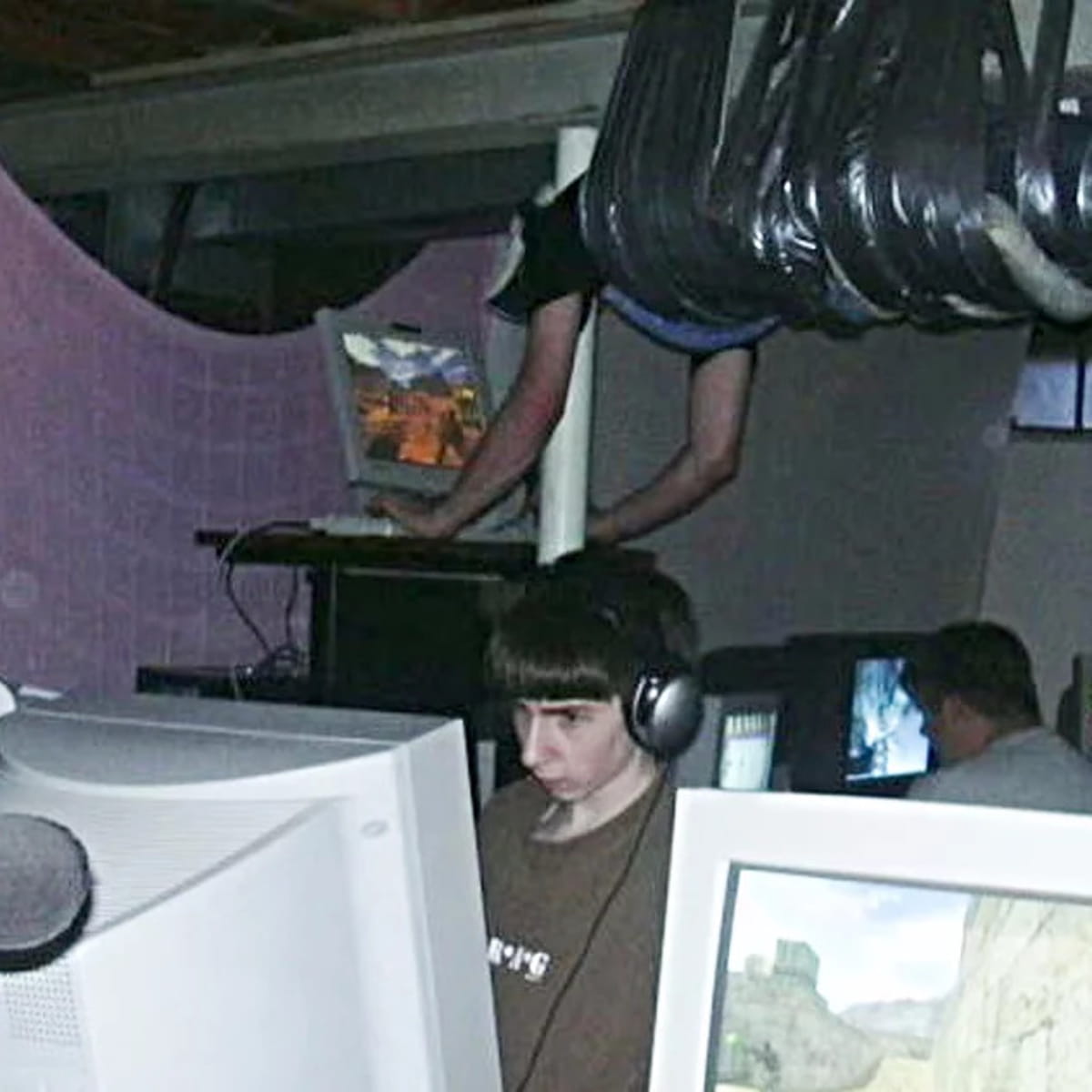 photo of a lan party, a person is duct taped to the ceiling in the background, leaning down to use a raised desktop computer