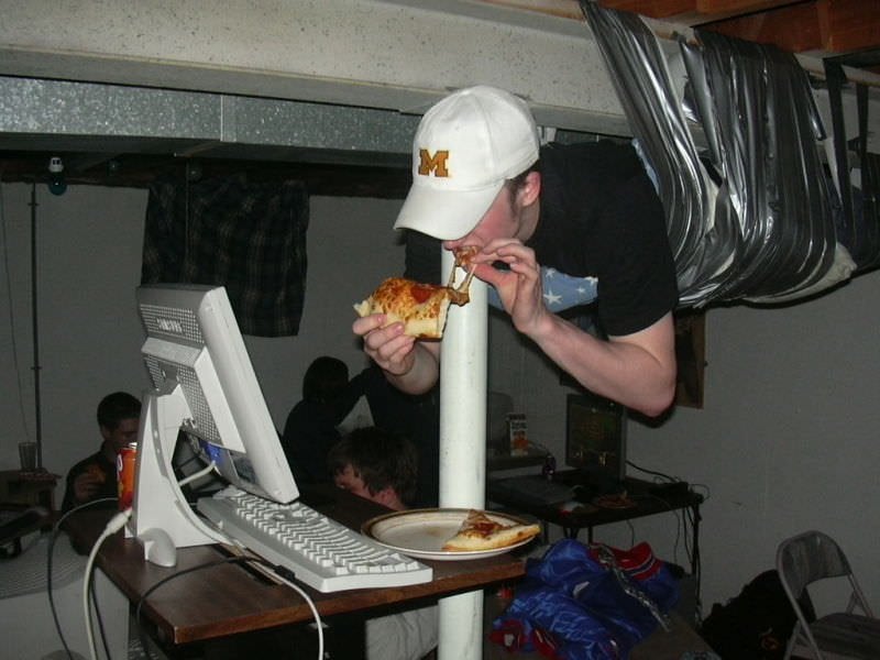 photo of a lan party, a person is duct taped to the ceiling, eating pizza from a tall computer desk